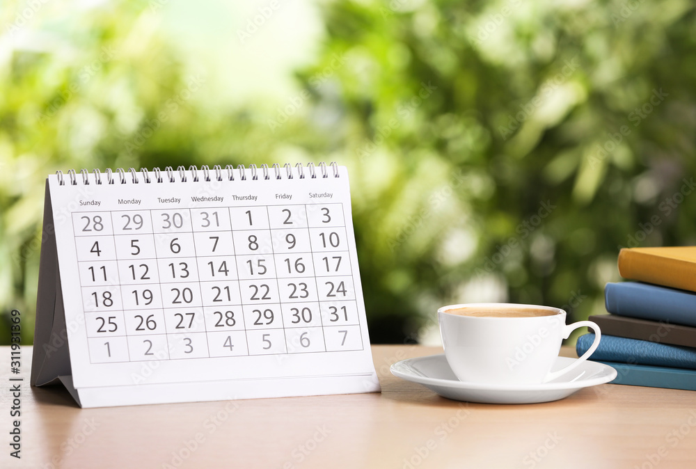Calendar and cup of coffee on wooden table against blurred background