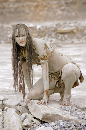 A young woman is  dressed as a neanderthal warrior.  She is covered with mud, filth and dirt and is seen in  a stone quarry area surrounding.
