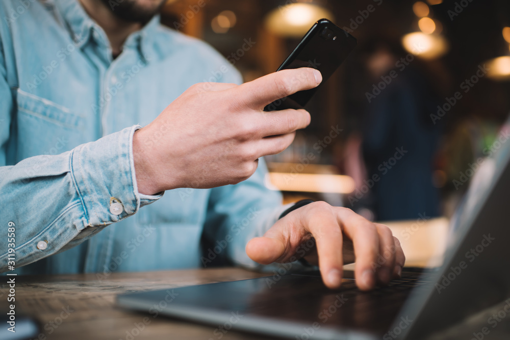 Faceless male typing on laptop while browsing mobile in workspace