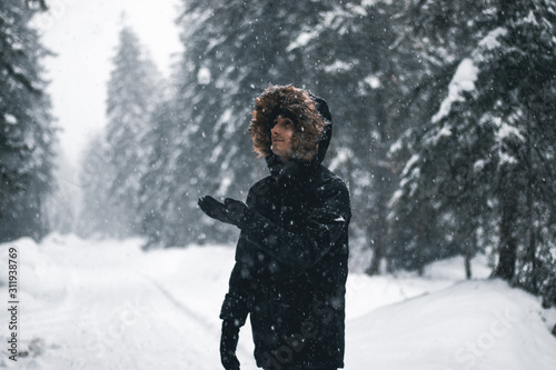 Heavy Snow Falling. A Guy Wearing A Black Coat With A Fur Hood And Holding His Hand Out For The Snow, Switzerland.