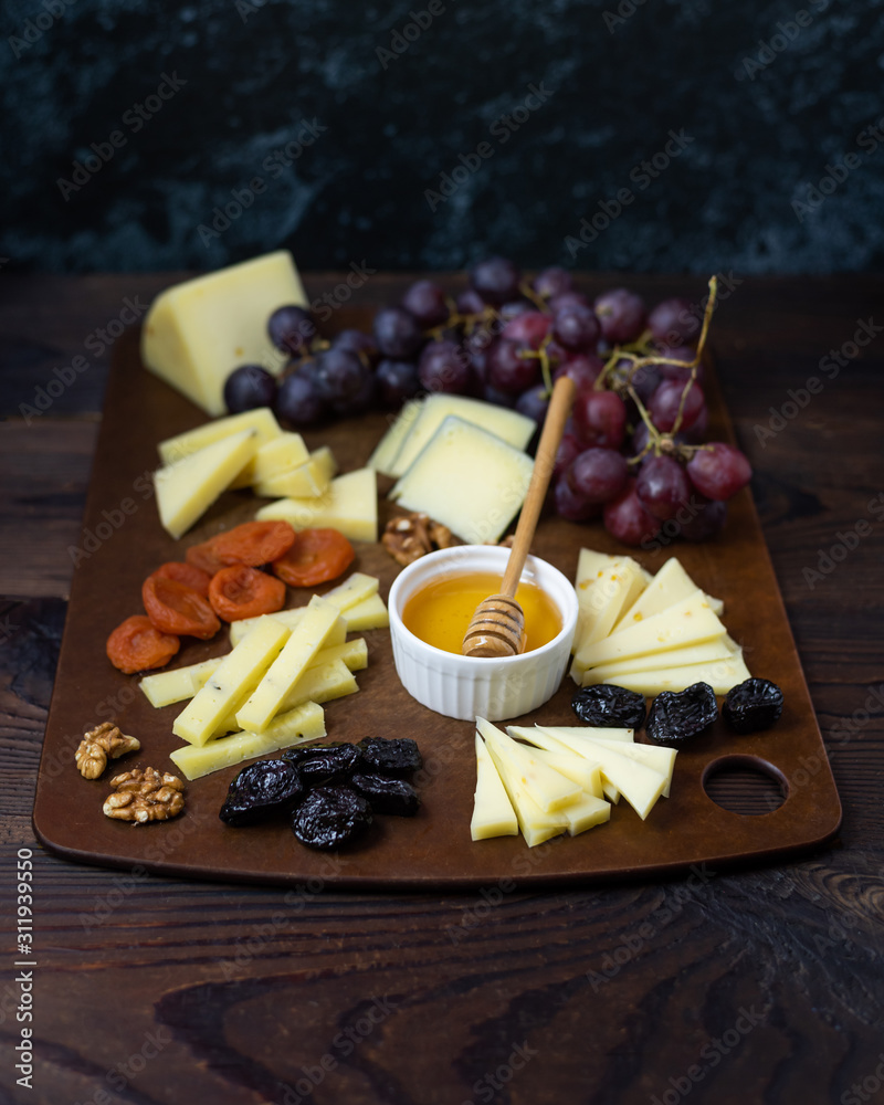 A large board with sliced different types of cheeses with honey, grapes, nuts and dried fruits. A set of cheese plates for a party and guests on a dark background.