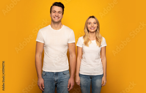 That’s us. Close-up photo of a beautiful couple, who are posing in white t-shirts on a yellow background with their arms down, looking in the camera and smiling.