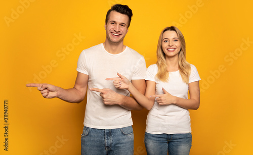 In the same direction. A beautiful couple is posing on a yellow background, wearing white t-shirts, pointing to the left with their hands and smiling at the camera.