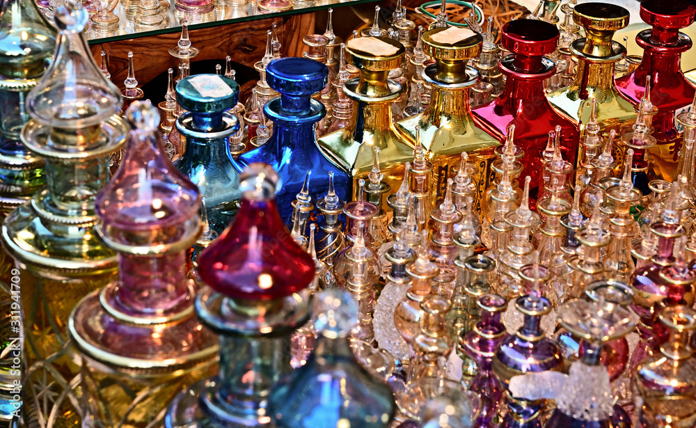 Close together standing richly decorated and colored perfume flacons made of glass in the light of the setting sun. The focus lies on the great flacons on the right side.