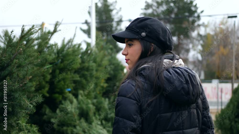 A hispanic woman shopping for a seasonal holiday Christmas tree decoration on a lot with many species of festive trees. A hispanic woman shopping for a seasonal holiday Christmas tree decoration on a lot with many species of festive trees.