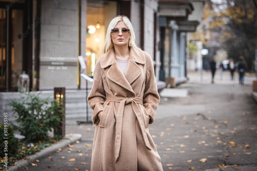 Fototapeta premium Young beautiful blond woman in an autumn coat stands on a city street. Soft focus. Fashionable concept.