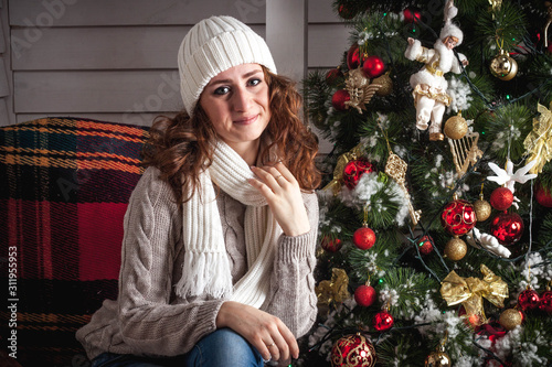 A red-haired girl in a white knitted hat and a knitted sweater sits in a chair on the porch near the Christmas tree, decorated in Christmas style. Winter