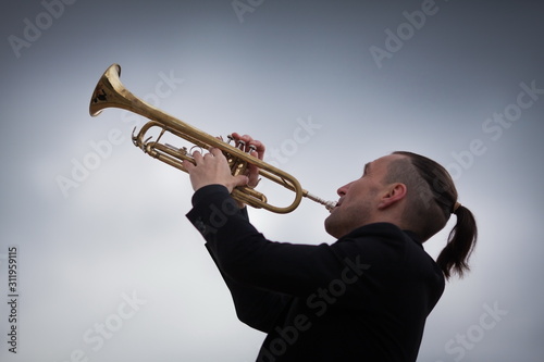 young trumpeter playing his musical instrument against background of dar grey gloomy sky with clouds