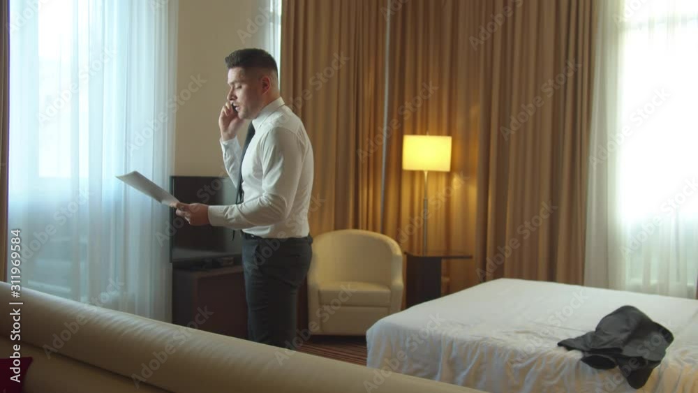 Man with documents in hands walk in room in hotel, talk on phone very emotionally Man with documents in hands walk in room in hotel, talk on phone very emotionally