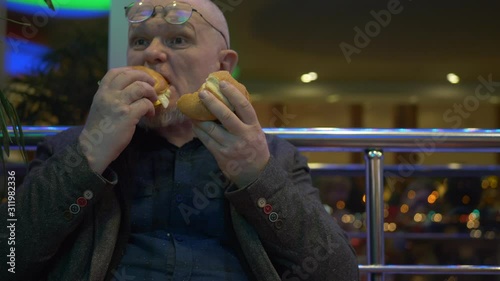 elderly pensioner in fashionable jacket and glasses with gray beard bites two hamburgers in turn. man is very hungry and angry