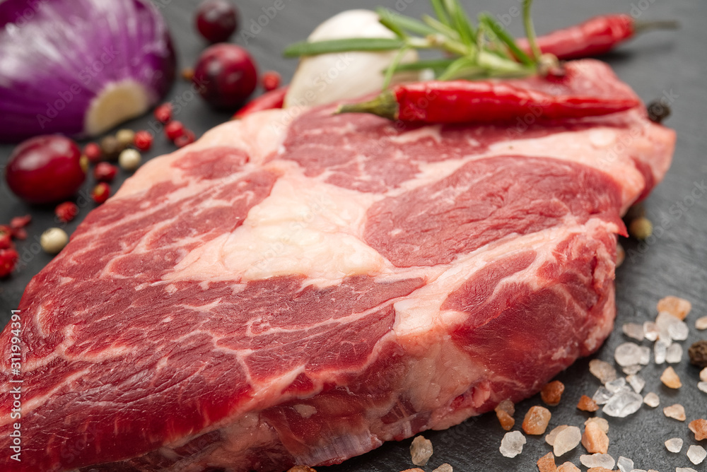Raw rib eye steak with spices and vegetables. Ingredients for restaurant meal. Fresh meat, salt, rosemary, thyme, chilli, cherry tomatoes, garlic on black stone. Food background. Raw rib eye steak with spices and vegetables. Ingredients for restaurant meal. Fresh meat, salt, rosemary, thyme, chilli, cherry tomatoes, garlic on black stone. Food background.