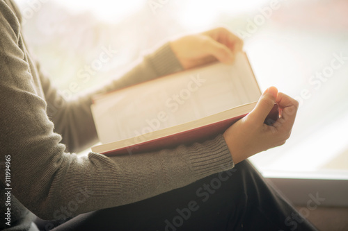 Close-up of an Asian woman reading an old book in the corner of a university library. With golden light shining through the glass of the room