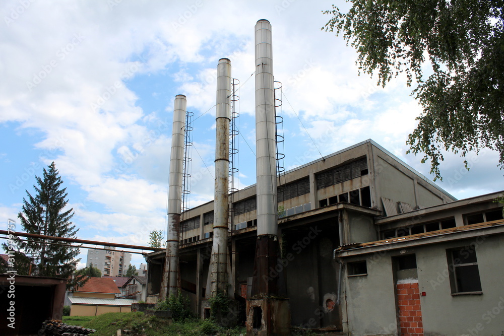 Abandoned old industrial complex building with destroyed backyard wall next to three tall metal chimneys surrounded with family houses and apartment buildings on cloudy blue sky background Abandoned old industrial complex building with destroyed backyard wall next to three tall metal chimneys surrounded with family houses and apartment buildings on cloudy blue sky background