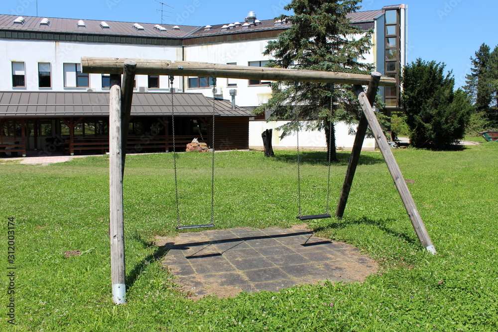 Fototapeta premium Dilapidated old wooden retro vintage outdoor public playground equipment in shape of swing with two seats in abandoned office building backyard surrounded with grass and trees