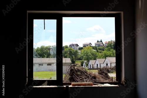 Large and small windows with broken and missing glass at abandoned house overlooking piles of cut down trees and military complex hangars surrounded with dense trees and family houses on warm sunny