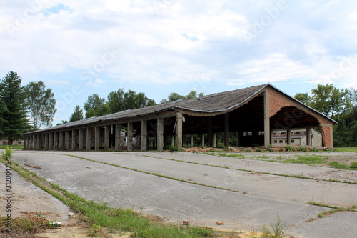 Large long hangar building with missing destroyed red brick support walls and unusual bent roof surrounded with paved area mixed with grass and trees at abandoned military complex on cloudy blue sky b © hecos