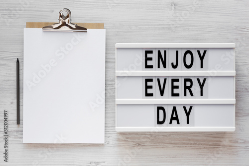 'Enjoy every day' words on a lightbox, clipboard with blank sheet of paper on a white wooden surface, top view. Overhead, from above, flat lay. photo