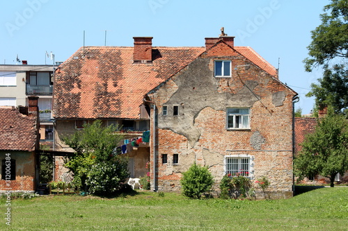 Old red brick suburban family house with cracked facade and windows with dilapidated wooden frames surrounded with grass and trees on clear blue sky background