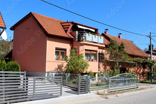 Renovated small suburban family house with new dark pink facade and top balcony filled with flowers surrounded with garden plants and metal fence next to other houses and paved street on warm sunny
