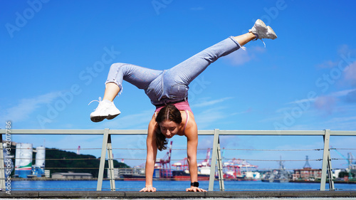 a young athletic girl equilibrist performs a stand on one arm on a sunny day. circus performer on the street. girl gymnast in life style clothes on the observation deck in the port. flexibility girl