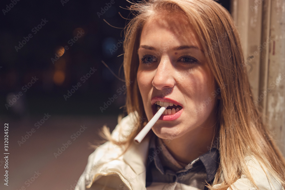 Fototapeta premium Close up portrait of young caucasian woman on the street at night holding cigarette in her mouth with teeth looking mean and furious mad
