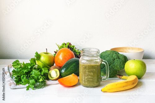 Detox green smoothie in a glass jar with ingredients: apples, kiwi, avocado, banana, persimmon, lettuce, broccoli, dill and parsley on white wooden table. Healthy raw vegan food concept.