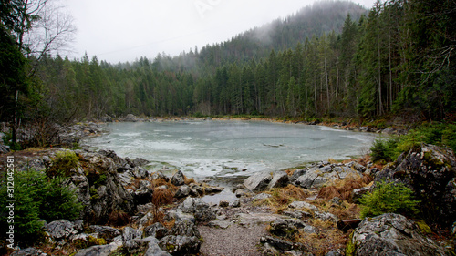 Walk around the Eibsea (Eibsee) near the Zugspitze (highest mountain in Germany). Rainy and foggy weather. Frozen side-sea surrounded with green fir trees and grey stones and rock fragments.