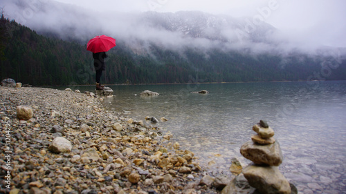 Walk around the Eibsea (Eibsee) near Zugspitze (highest mountain in Germany). Sea surrounded with green fir trees, grey stones and rock fragments. Foggy weather. Wanderer with red umbrella. Stone Art.