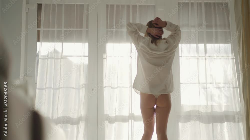 Back view of Caucasian girl in lace panties and white shirt standing in front of big window and stretching. Young attractive woman resting in bedroom in early morning. Leisure, lifestyle, silence.