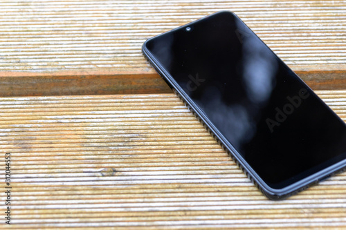 Schwarzes Smartphone Phablet liegt auf einem Holztisch im Garten und zeigt mit copy space mobile Erreichbarkeit und Nutzen in Freizeit und draußen sein