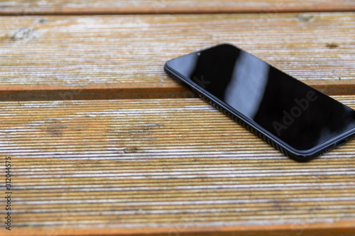 Schwarzes Smartphone Phablet liegt auf einem Holztisch im Garten und zeigt mit copy space mobile Erreichbarkeit und Nutzen in Freizeit und draußen sein