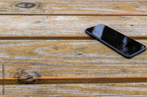 Schwarzes Smartphone Phablet liegt auf einem Holztisch im Garten und zeigt mit copy space mobile Erreichbarkeit und Nutzen in Freizeit und draußen sein