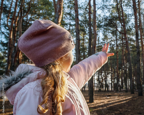 A cute girl in a pink jacket stands in the forest, raises her hand and tries to close herself from the bright sun.