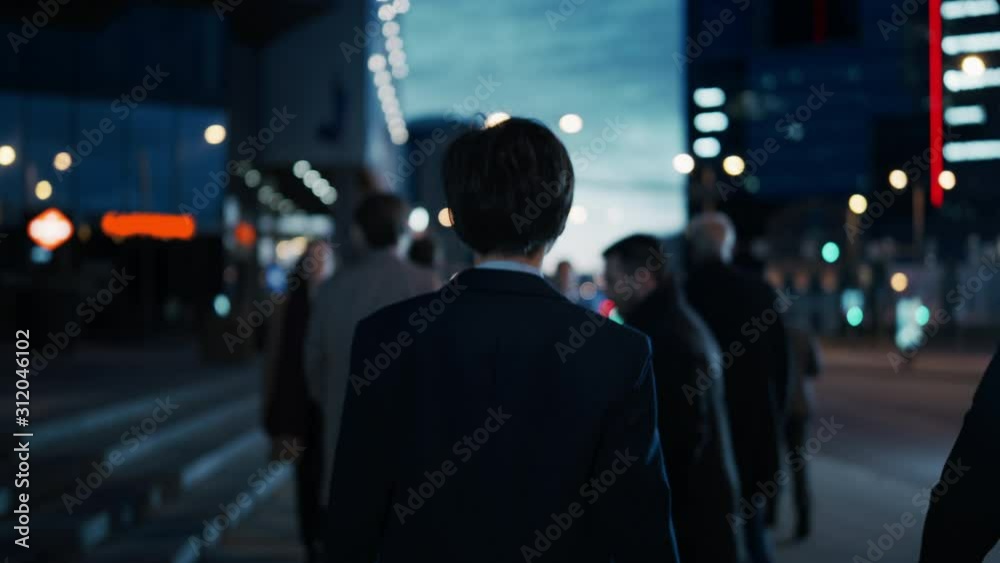 Businessman in a Suit Walking on the Street. Other Office People Commute to Work or Home. It's Early Morning or Late in the Evening. Footage from the Rear. Urban City Lights in the Background. Businessman in a Suit Walking on the Street. Other Office People Commute to Work or Home. It's Early Morning or Late in the Evening. Footage from the Rear. Urban City Lights in the Background.