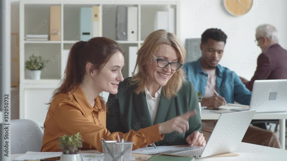 Waist-up shot of young Caucasian female office worker sitting at desk together with mature manager, showing her something on laptop screen and explaining while woman is watching and smiling Waist-up shot of young Caucasian female office worker sitting at desk together with mature manager, showing her something on laptop screen and explaining while woman is watching and smiling