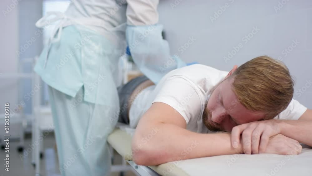 Tracking shot of Caucasian man lying on stomach on medical couch in hospital while nurse in uniform giving him injection into buttock