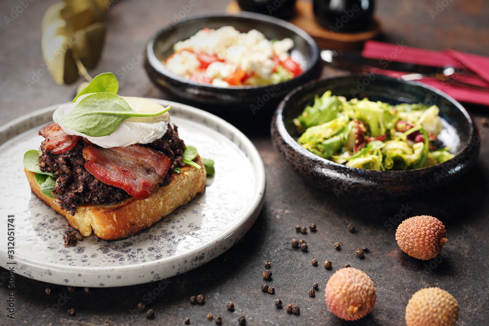 Toast with black pudding, poached egg, bacon and mayonnaise sauce. Food, ready dish on a dark background. Suggestion of serving the dish. Toast with black pudding, poached egg, bacon and mayonnaise sauce. Food, ready dish on a dark background. Suggestion of serving the dish.