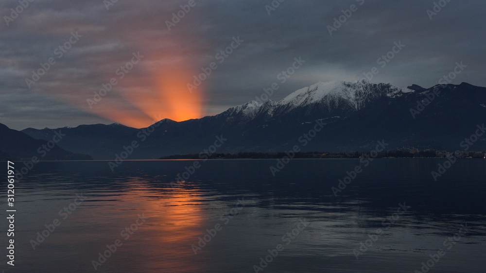 Tramonto sul lago con sole che tramonta e crea cono di luce emergente dalla montagna
