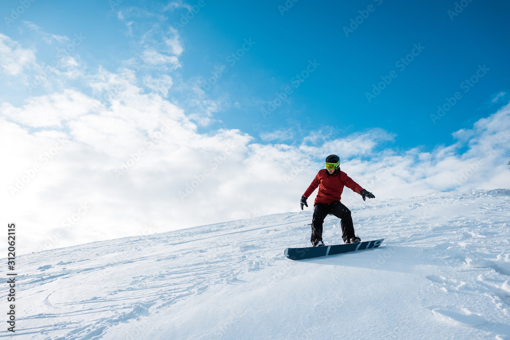 athletic snowboarder in helmet riding on slope against blue sky with clouds
