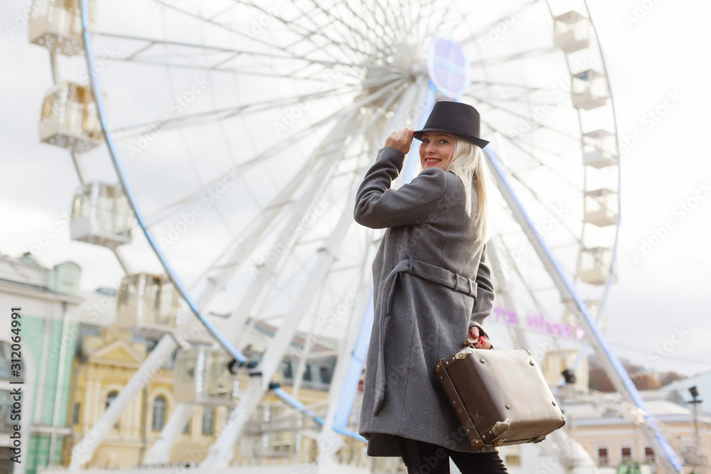Attractive blonde girl in casual clothing, wearing a coat, holding a brown retro suitcase in his hands, background of the ferris wheel Attractive blonde girl in casual clothing, wearing a coat, holding a brown retro suitcase in his hands, background of the ferris wheel