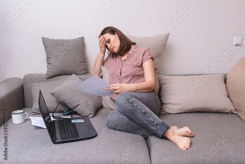 white girl in jeans is sitting on the couch next to a laptop and reading a document with a tired expression on her face,