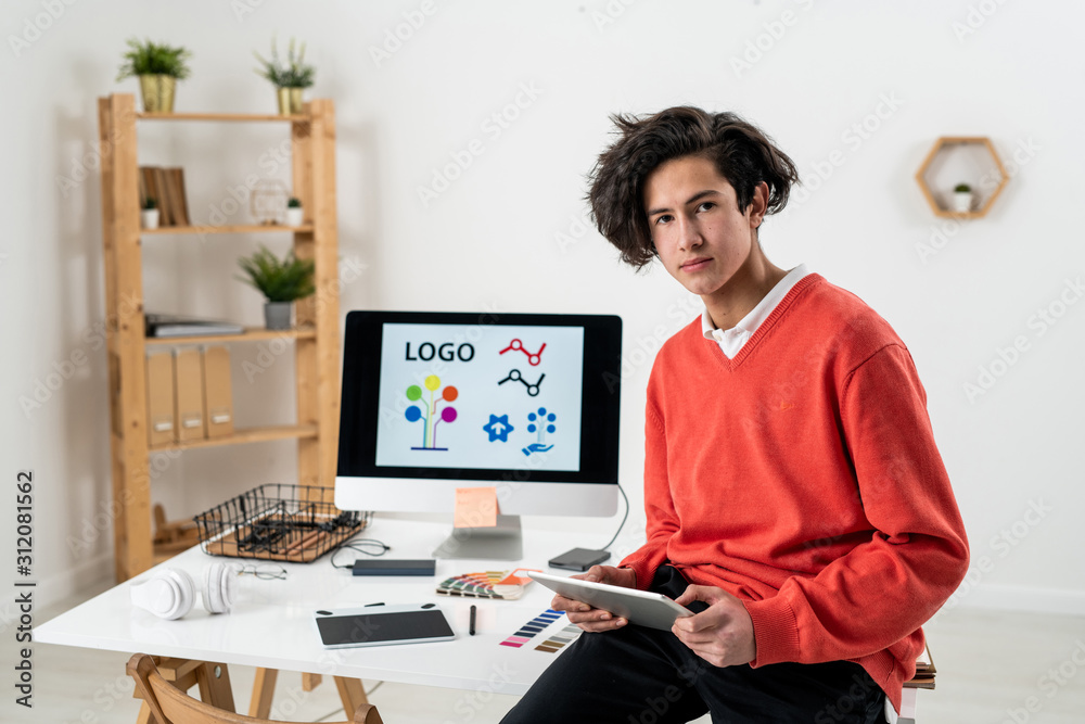 Young serious freelance designer with touchpad sitting on desk at home office Young serious freelance designer with touchpad sitting on desk at home office