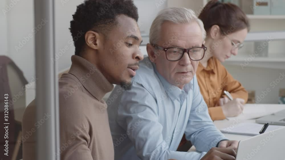 Chest-up shot of senior Caucasian man pointing at laptop screen and listening attentively while young Afro-American student giving detailed explanations of project Chest-up shot of senior Caucasian man pointing at laptop screen and listening attentively while young Afro-American student giving detailed explanations of project