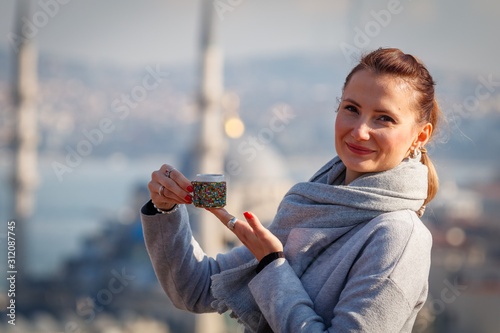 Beautiful woman stands at the rooftop an looking to the camera with colorful cup of tea with, New mosque on the background, Istanbul, Turkey
