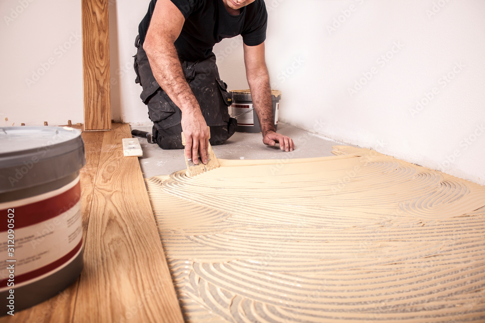 Handwerker verlegt Parkettboden und streicht den Estrich mit Kleber ein und  schlägt die Dielen mit dem Hammer und dem Schlagklotz zusammen Stock-Foto |  Adobe Stock