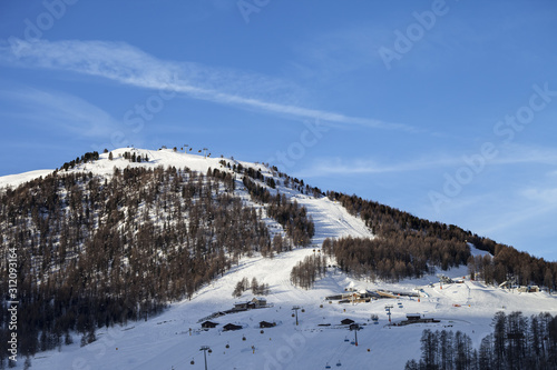 Snowy ski slope with ski-lifts, traces and forest in high winter mountains at sunny morning