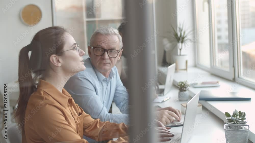 Waist-up shot of excited Caucasian female intern talking to attentive senior male mentor in office, who is listening and nodding, and black man working on laptop and writing in notebook in background Waist-up shot of excited Caucasian female intern talking to attentive senior male mentor in office, who is listening and nodding, and black man working on laptop and writing in notebook in background