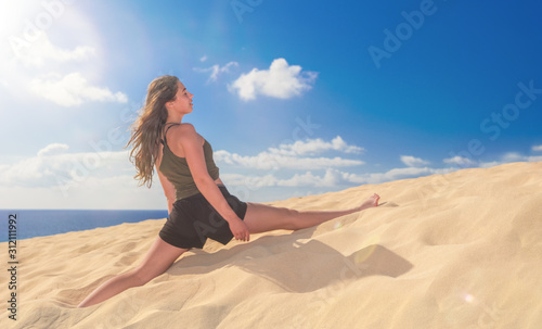 ein junges Mädchen sitzt im Spagat auf einer Düne im gelben Sand vor blauem Himmel und macht Yoga