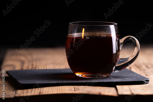  a transparent glass cup with hot tea stands on an old wooden surface on a black background with a place for inscription