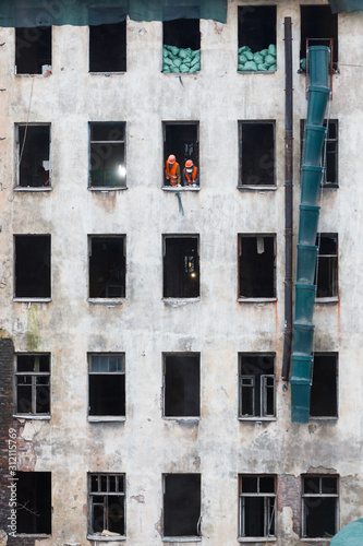 Dismantling or reconstruction with a complete replacement of all communications inside of the old house, construction workers in orange vests and hardhat standing by the window. Construction site. 